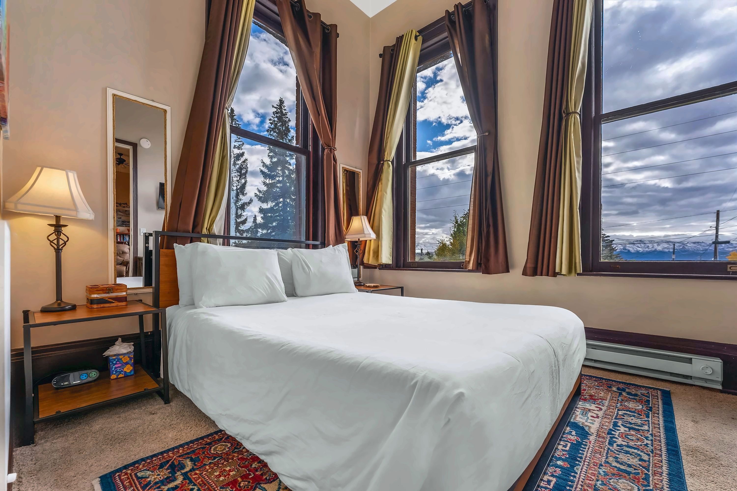 A hotel room with a bed, a nightstand, a lamp, a mirror, and a view of trees through large windows.
