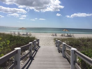 Ein hölzerner Steg führt zu einem Strand mit Liegestühlen und Sonnenschirmen.