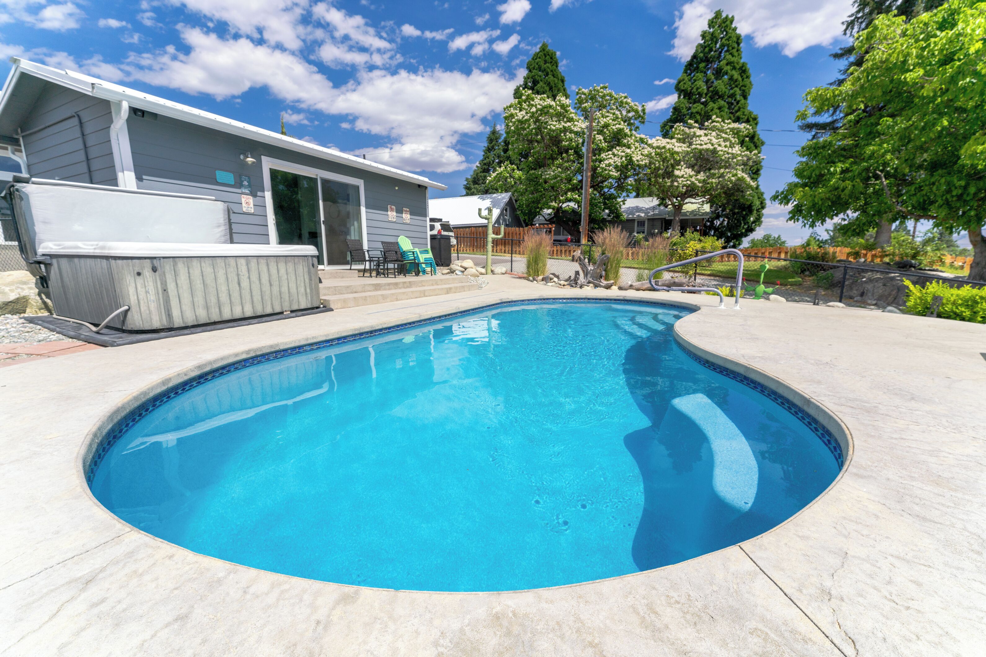 A swimming pool with a hot tub, a covered patio, and a view of a residential area with trees and a house.