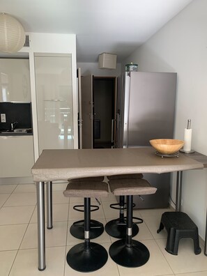 A modern kitchen with a large table, stools, a refrigerator, and a door leading to another room.