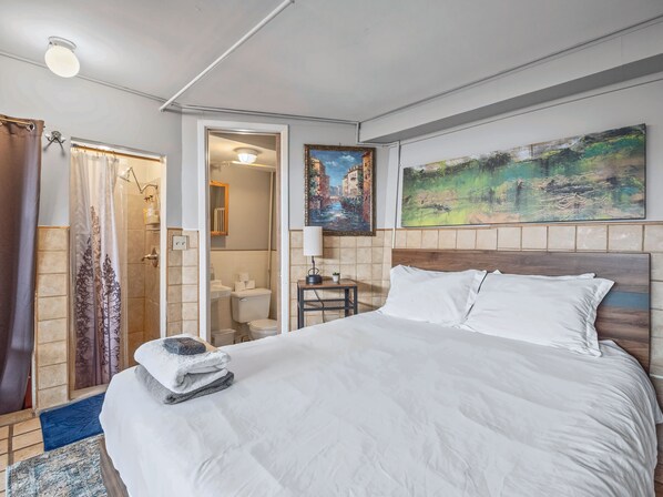 A hotel room with a bed, a shower area, a bathroom, and a painting on the wall.