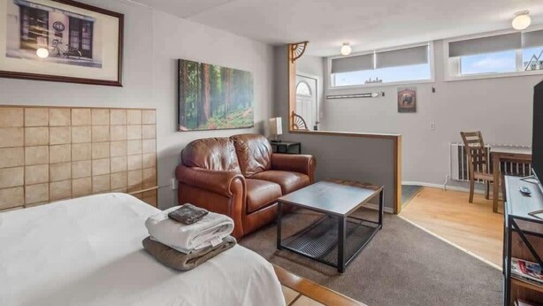 A modern hotel room with a bed, a brown leather sofa, a small table, a painting, and a view of the city through large windows. - 1BR Cozy Downtown Studio Condo: Hiking Nearby (Leadville)