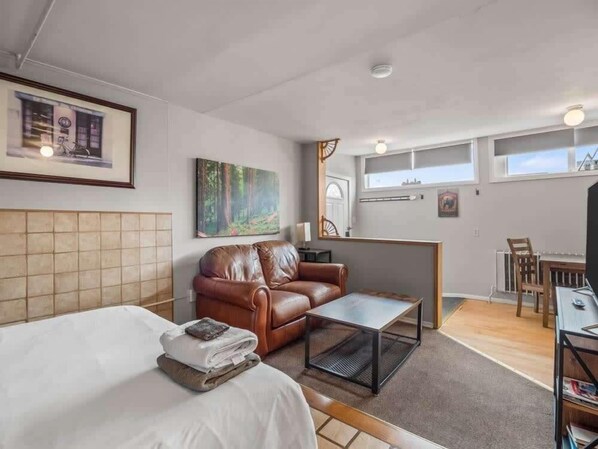 A modern hotel room with a bed, a brown leather sofa, a small table, a painting, and a view of the city through large windows.
