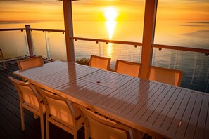 Une terrasse en bois avec une table et des chaises, donnant sur une étendue d’eau au coucher du soleil.