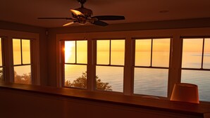 Una habitación con ventilador de techo, ventanas amplias que dan a una vista del atardecer sobre el agua, y una lámpara en el alféizar.
