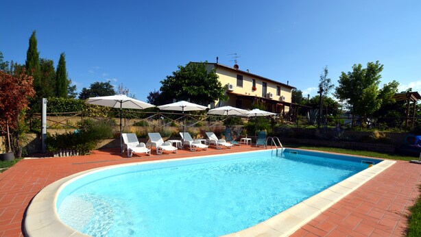 A swimming pool with lounge chairs and umbrellas, surrounded by a brick patio and a building in the background.