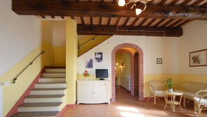 A staircase leading to a landing with a TV on a cabinet, a sofa, and a table with chairs in a sunlit room with exposed wooden beams.