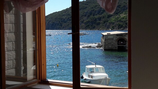 A view through a window of a coastal area with a boat docked near a stone building, surrounded by water and hills.