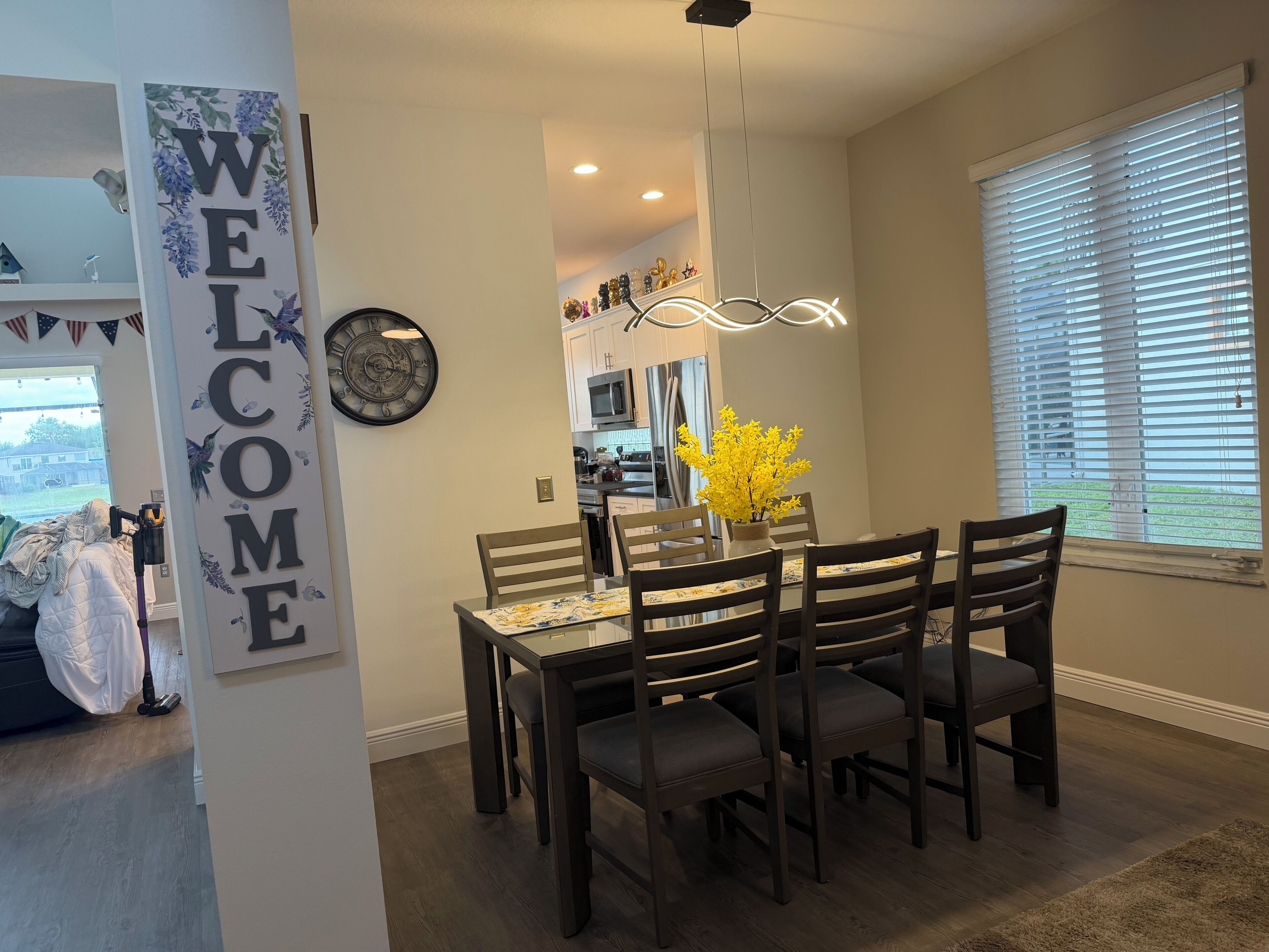 A dining area with a 'WELCOME' sign, a clock, and a vase with yellow flowers.