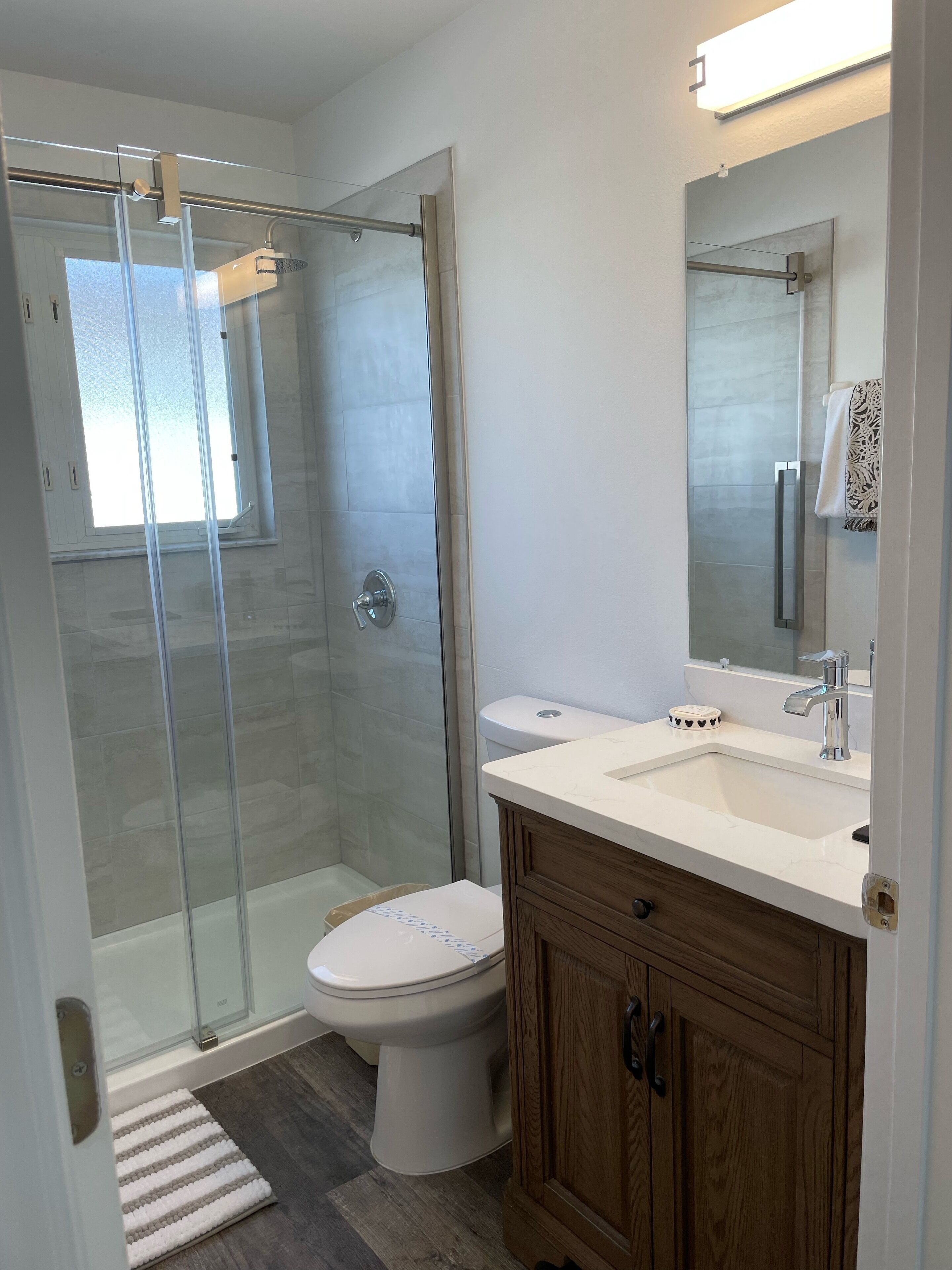 Une salle de bain moderne avec une douche à parois de verre, des toilettes blanches, un meuble-lavabo en bois et un grand miroir.