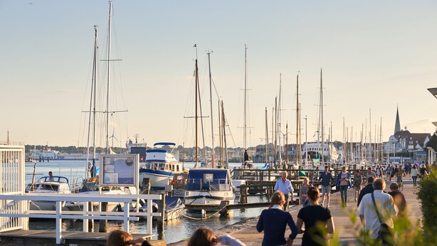 Un port de plaisance où sont amarrés de nombreux bateaux, une promenade piétonne et, au loin, le clocher d’une église.