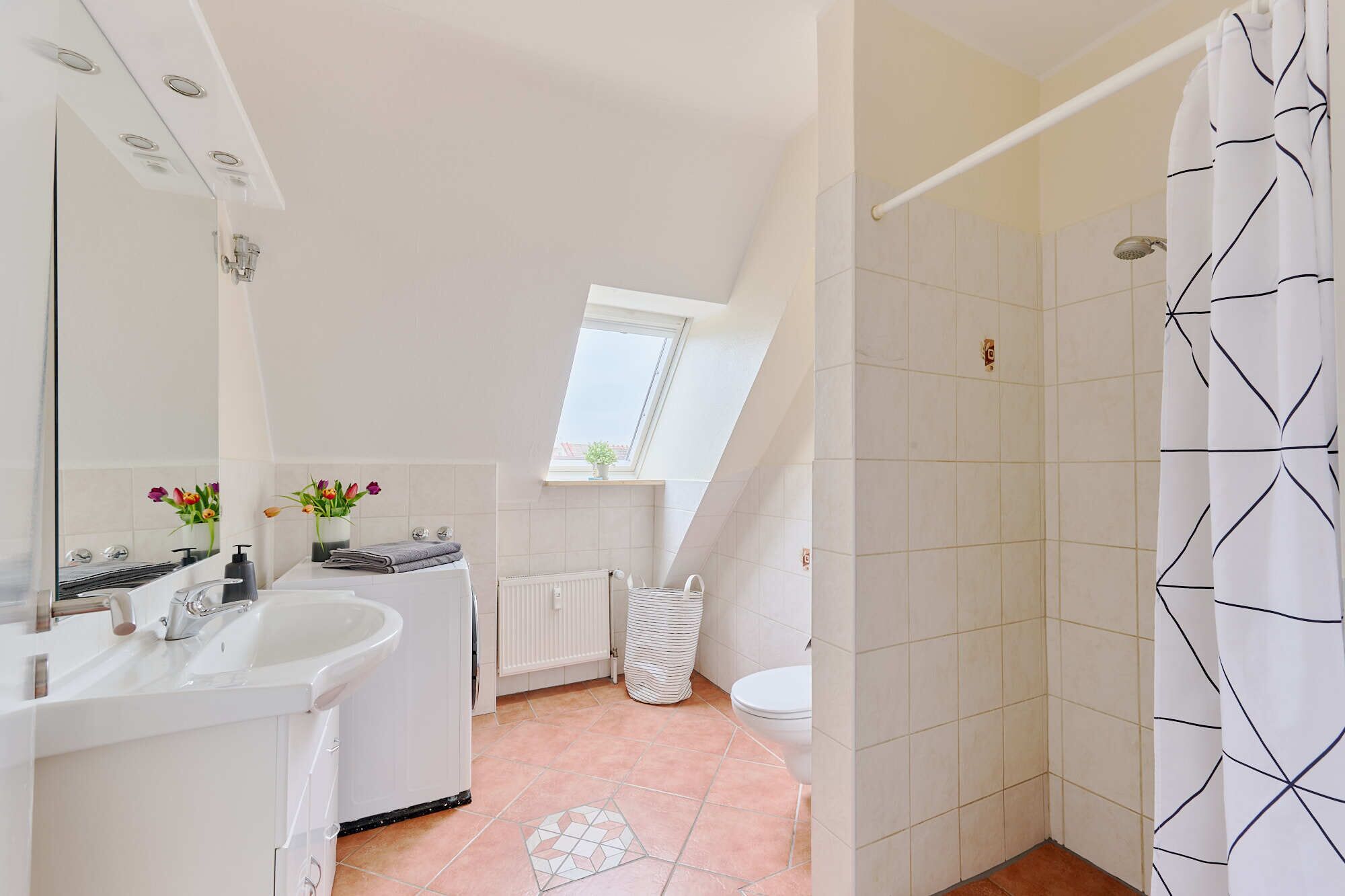 Ein modernes Badezimmer mit Oberlicht, weißen Fliesen, einer Duschvorhang mit geometrischem Muster und einem sichtbaren Heizkörper.