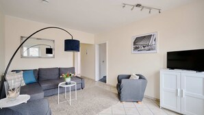 Un salon avec un canapé gris, une petite table ronde sur laquelle se trouve un vase, un lampadaire, un téléviseur posé sur un meuble et un tableau encadré accroché au mur.