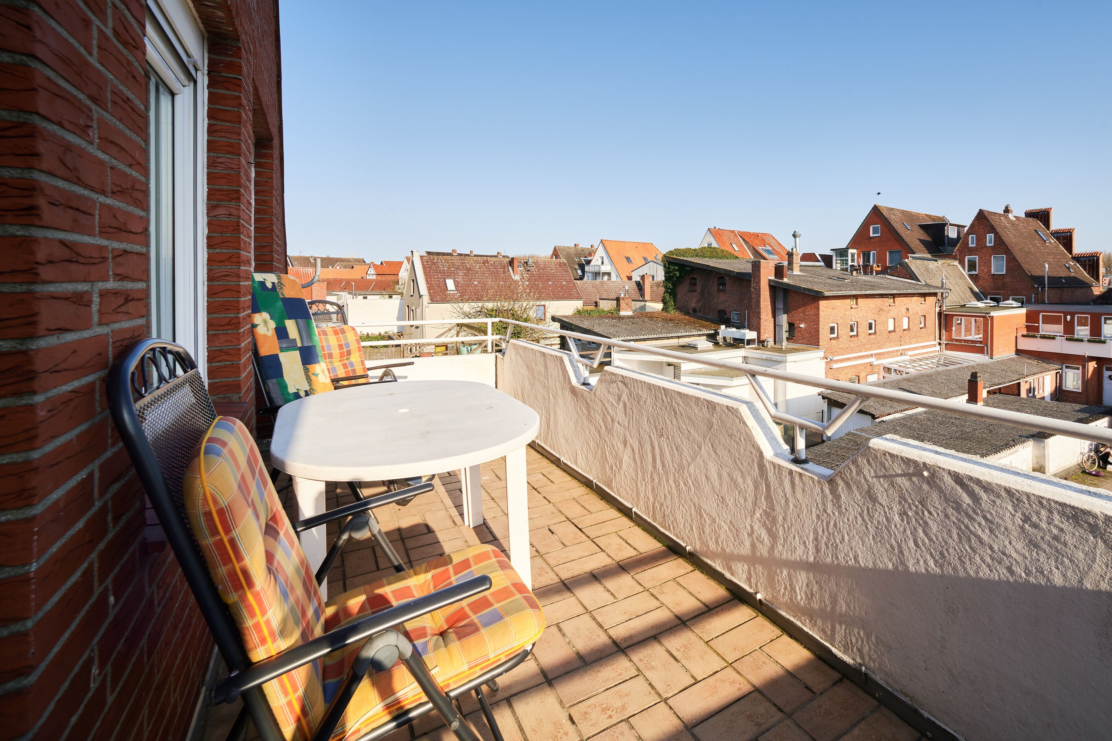 A balcony with a table and chairs, overlooking a residential area with brick houses.
