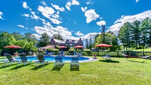 Área de piscina com espreguiçadeiras e guarda-sóis, cercada por uma vedação e árvores.
