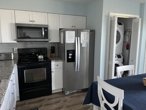 A compact kitchen with a microwave, stove, refrigerator, and a small dining area with a table and chairs. - CORAL SURF C-1 OCEANFRONT 2 BR & 2 BTH. PRIVATE 240V EV CHARGER & 110V OUTLET (Carolina Beach)
