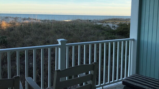 Una terraza de madera con barandilla blanca, con vistas a una playa con vegetación escasa y cielo despejado.