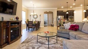 A modern living room with a flat-screen TV, a glass coffee table, a sofa, and a dining area with chairs and a chandelier. - Very spacious, private location with open area, good for family travel (Sugar Land)