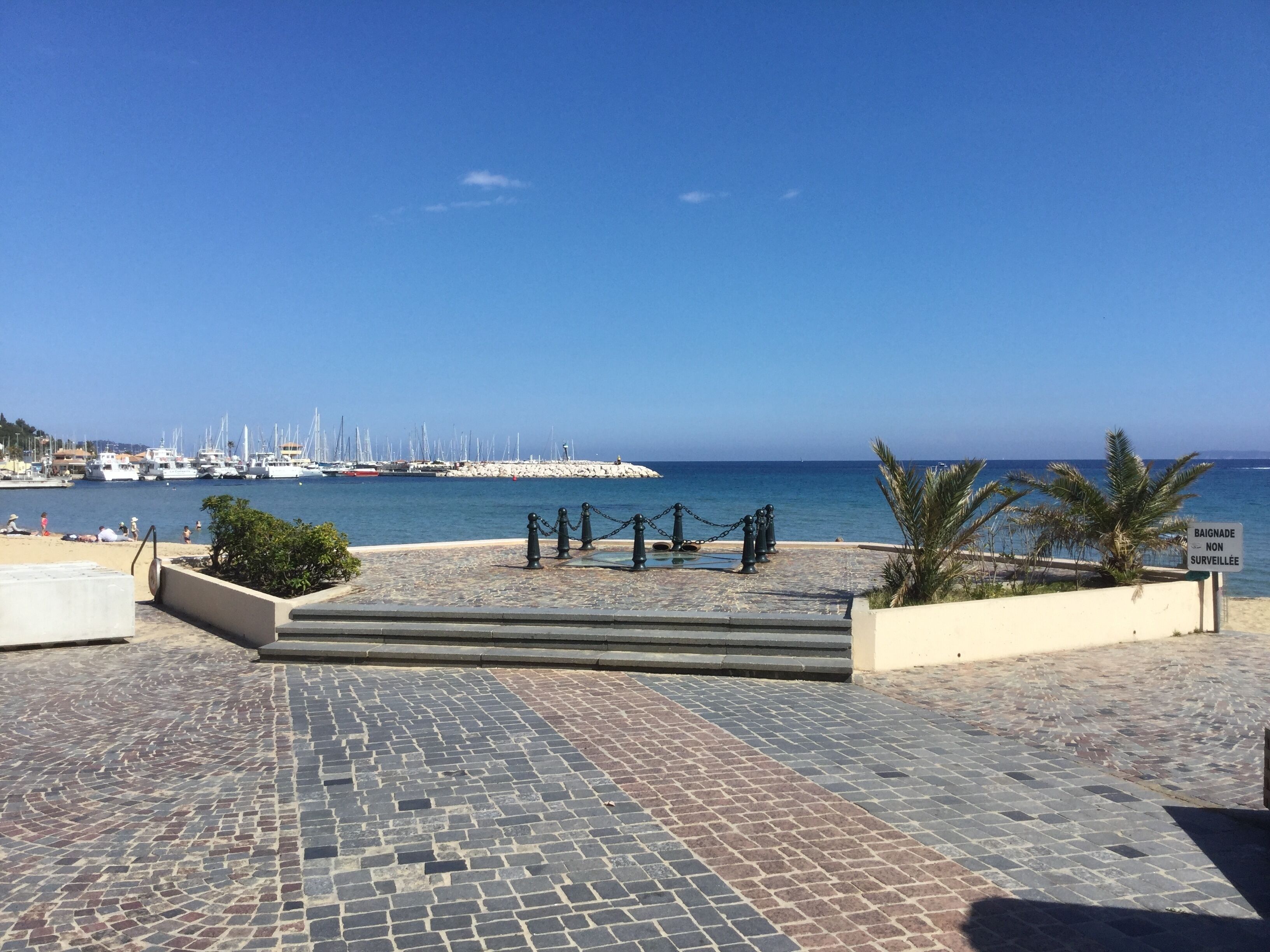 A paved walkway leading to a beach with a clear view of the sea and boats in the distance.