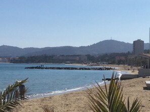 A beach with a clear view of the sea, sandy shore, and distant buildings. - Beautiful comfortable apartment 300m from the beach AU LAVANDOU (Le Lavandou)