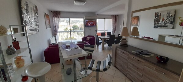 A modern hotel room with a dining area, a sofa, a desk with a computer, and a view of the city through large windows.