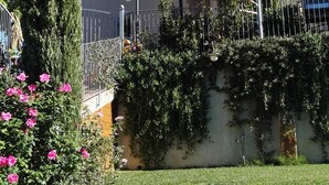 Une maison à deux étages avec un balcon, une cheminée et un jardin où poussent des fleurs roses.