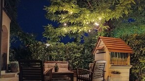 Un espace extérieur aménagé avec du mobilier en bois et une petite structure en bois, peut-être un kiosque, entouré de verdure et illuminé par des guirlandes lumineuses.