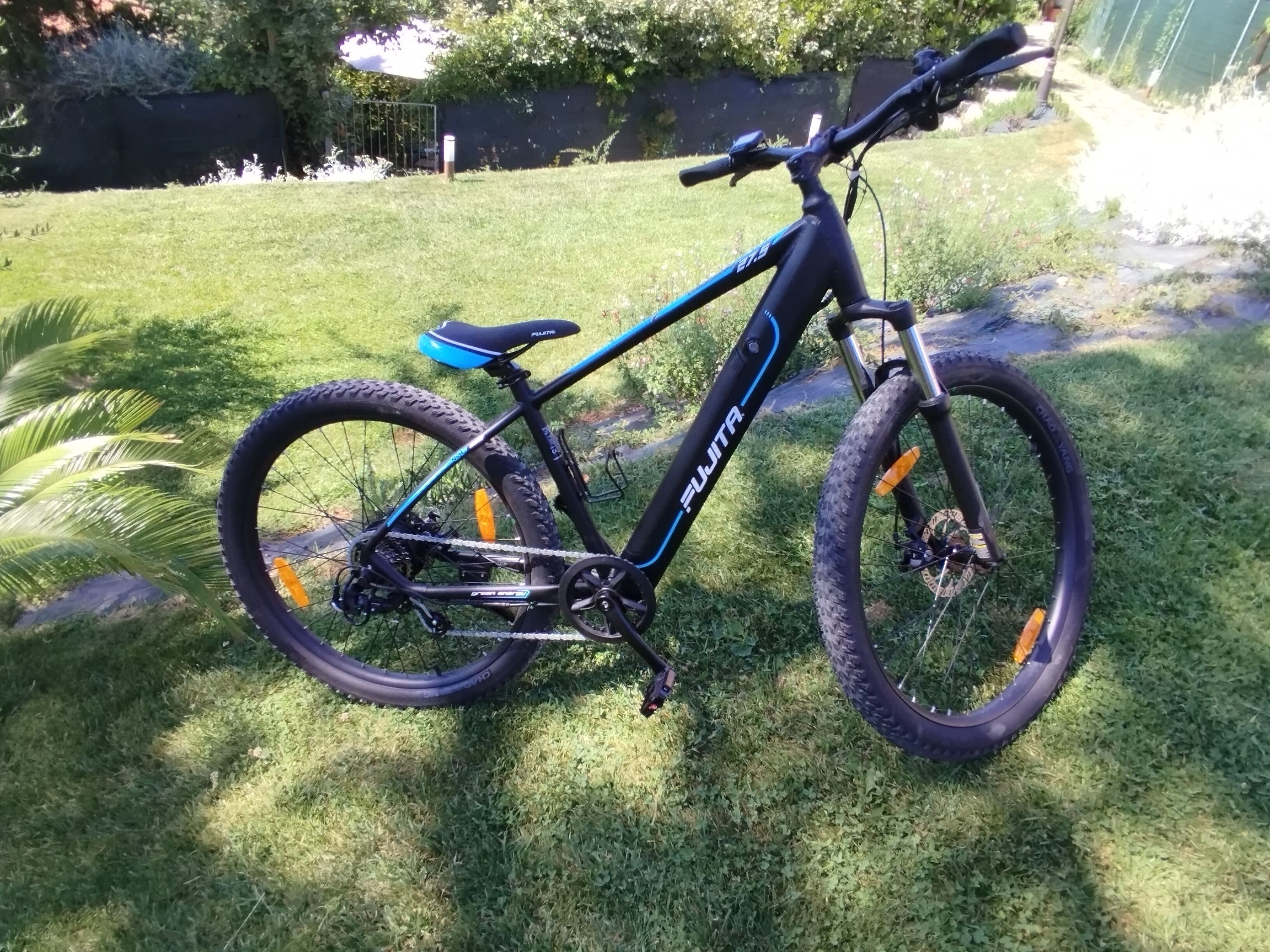 Un vélo tout-terrain avec un cadre noir et bleu, une selle et un porte-bagages arrière, garé sur l’herbe.