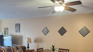 Uma área de refeições com mesa e cadeiras de madeira, um ventilador de teto, detalhes decorativos na parede e uma televisão ao fundo.