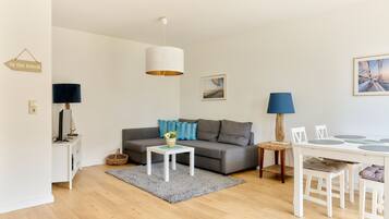 A modern living room with a grey sofa, a white coffee table, a dining table with chairs, a hanging light fixture, and a framed picture on the wall.