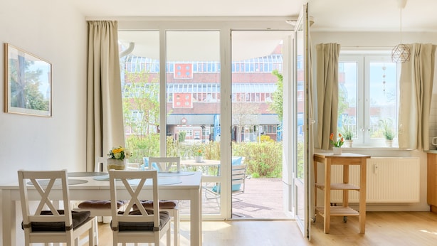 Une pièce lumineuse comprenant une table à manger, des chaises et une vue sur un immeuble à la façade rouge, visible à travers une porte vitrée.
