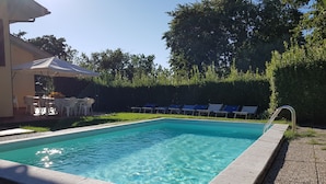 Une piscine rectangulaire à l’eau d’un bleu limpide, entourée d’une terrasse en galets. On y trouve des chaises longues et un espace abrité avec des tables et des chaises.