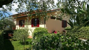 Une maison avec un toit en tuiles, des volets rouges et un jardin où poussent différentes plantes et fleurs.