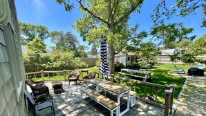 A deck with outdoor furniture, a picnic table, and a large tree providing shade. - Beautiful Home with Central A/C. Walk to the Beach, Large Yard (Dennis Port)