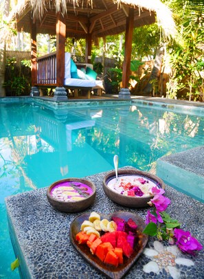 A pool area with a thatched-roof structure, lounge chairs, and a table with bowls of food and fruit. - Meno Island Villas (Gili Meno)