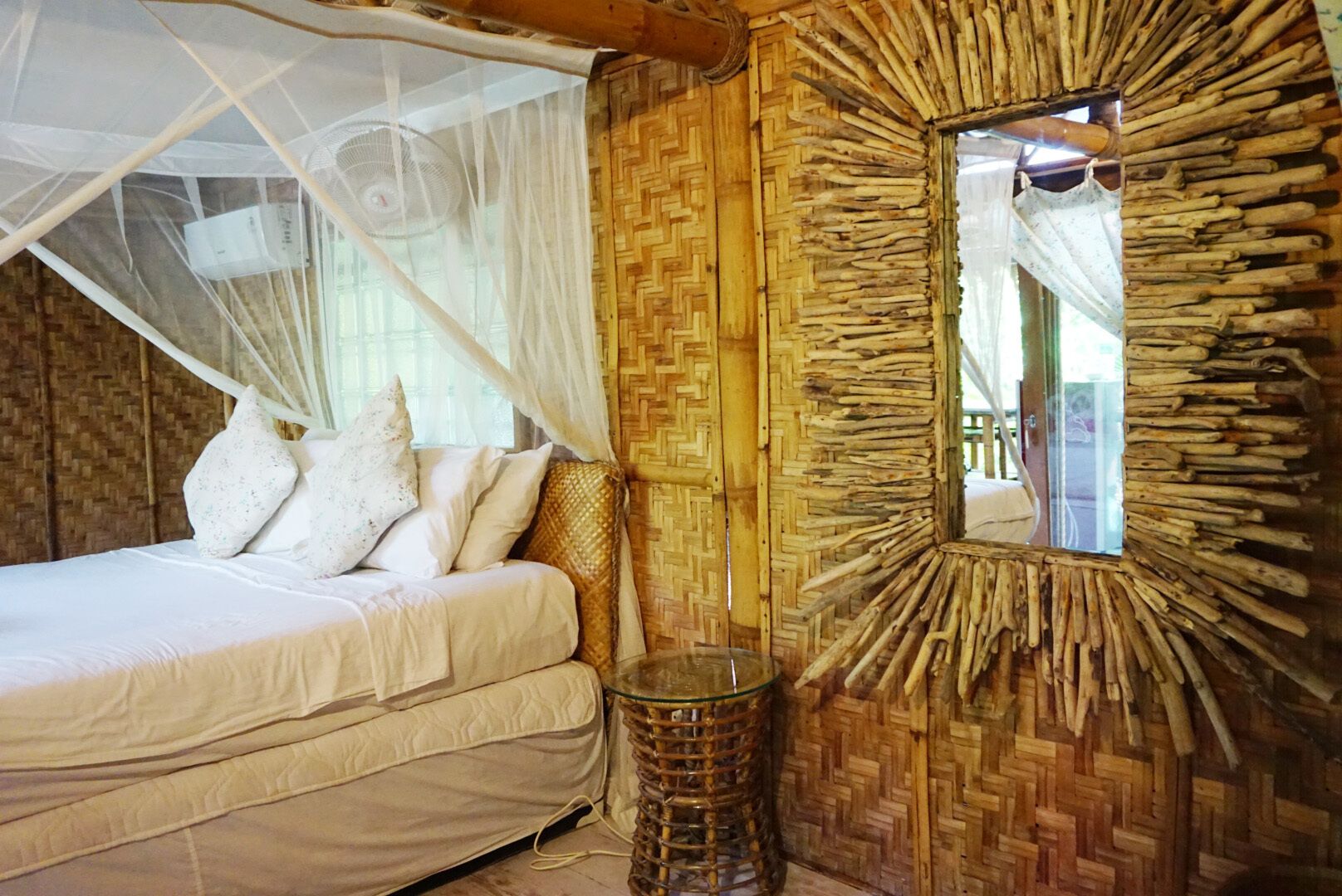 A bed with white bedding and a mosquito net, a woven wall, and a mirror with a wooden frame.