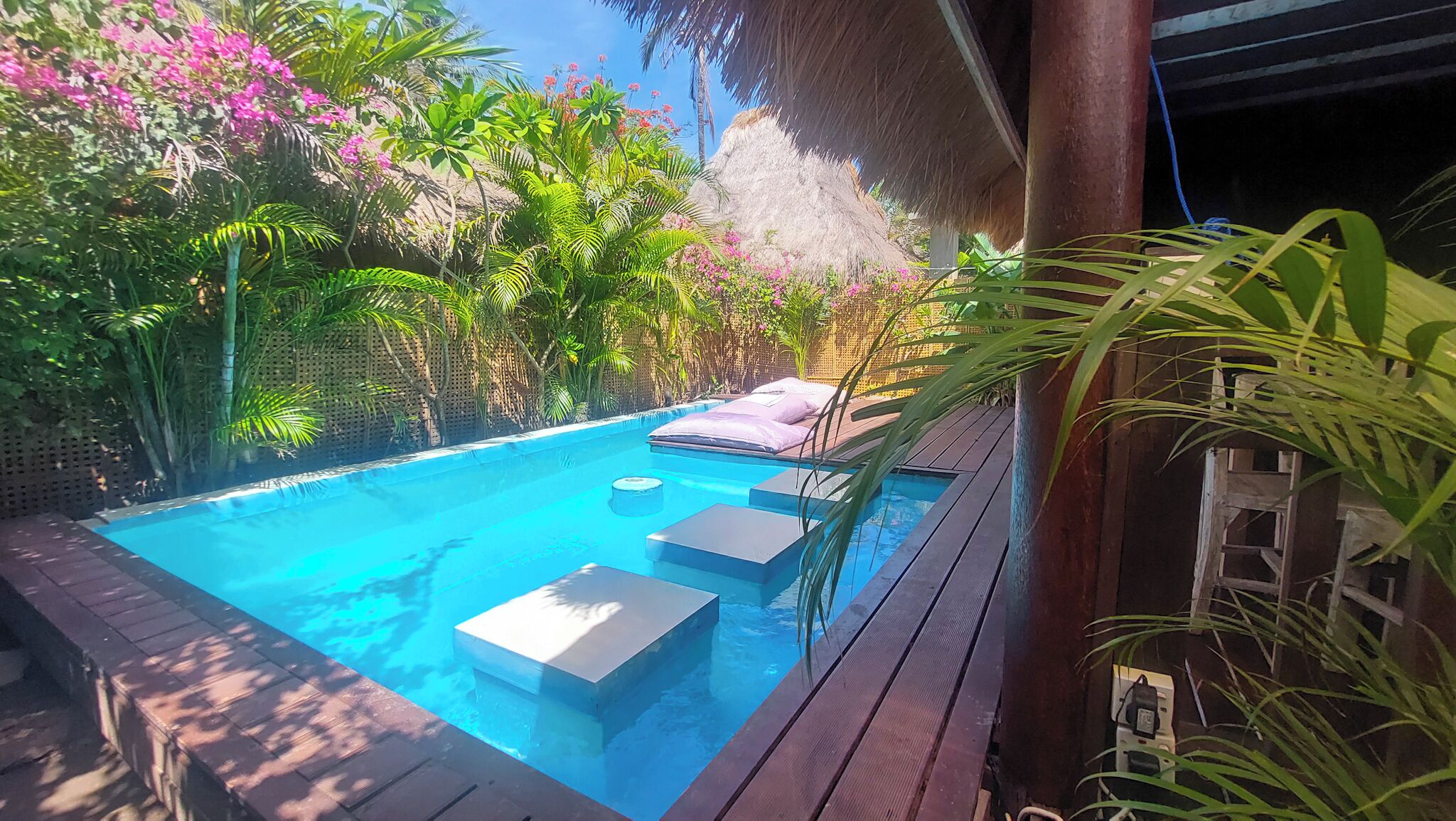 A swimming pool with steps leading into it, surrounded by lush greenery and a thatched roof structure.