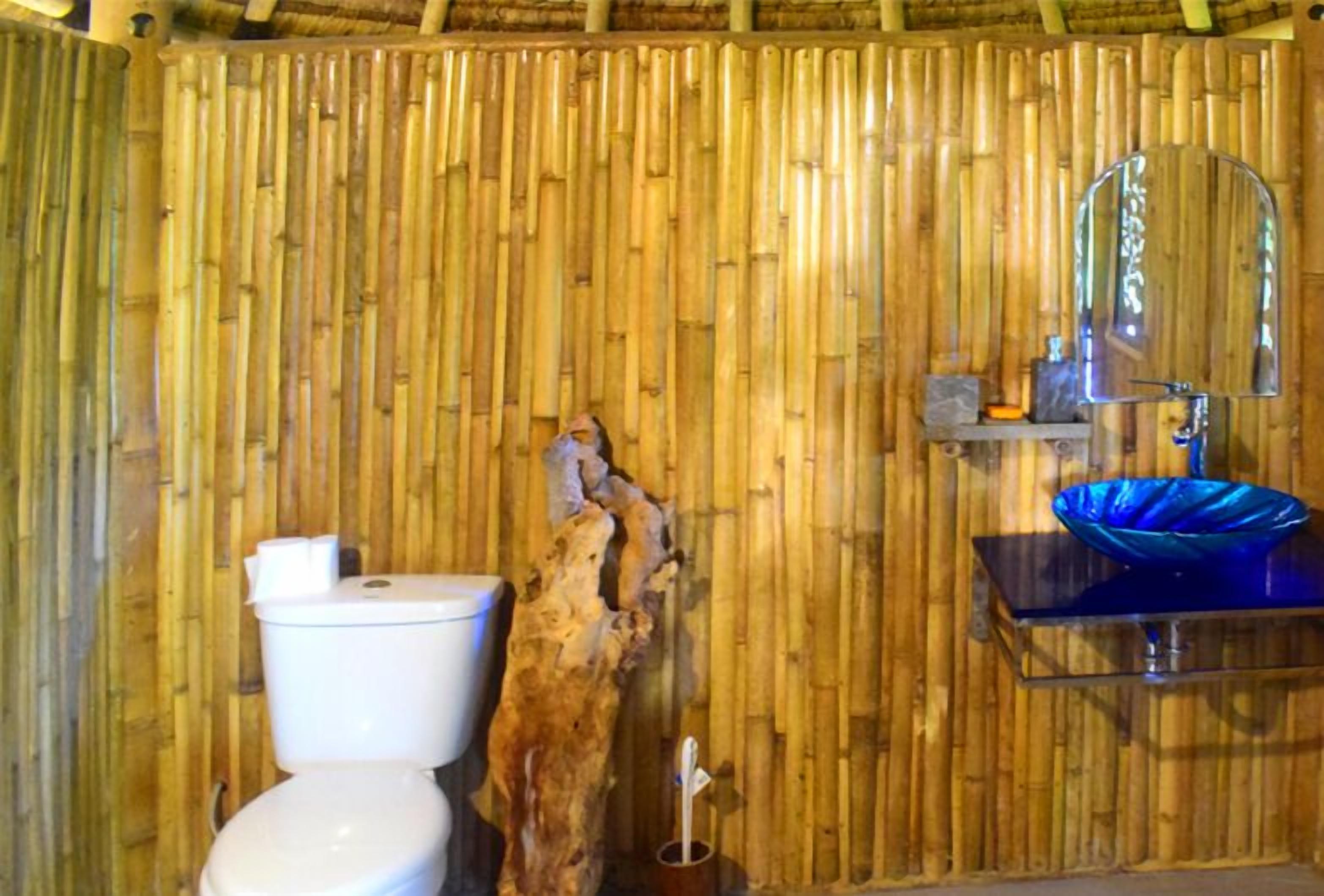Un baño con pared de bambú, inodoro blanco, lavabo azul y un espejo.