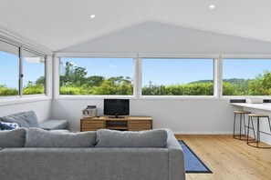A modern living room with a grey sofa, a TV on a wooden cabinet, and large windows offering a view of greenery. - Len's Place (Lorne)