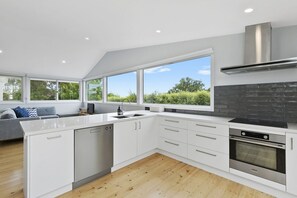A modern kitchen with white cabinetry, a built-in oven, and a large window overlooking greenery. - Len's Place (Lorne)
