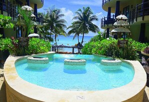 Une piscine dotée de trois bains à remous, entourée d’une végétation luxuriante et de palmiers, avec vue sur l’océan et un bâtiment en arrière-plan.