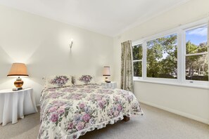 Ein Schlafzimmer mit Blumenoptik auf der Bettdecke, zwei Nachttischlampen, einem Nachttisch und einem Fenster mit Vorhängen.