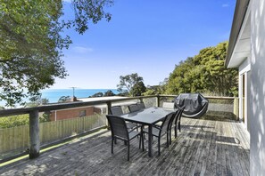 Eine Holzterrasse mit Tisch und Stühlen, einem Grill und Blick auf das Meer.
