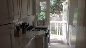 Een keuken met witte kasten, een fornuis en een koffiezetapparaat. Op de vloer ligt een klein vloerkleed en er is uitzicht op een balkon met een balustrade.