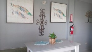 Un área de recepción con un mostrador blanco, dos cuadros enmarcados en la pared, una pieza decorativa de metal, una planta pequeña en maceta, un extintor y un adorno de árbol colgado en la pared.