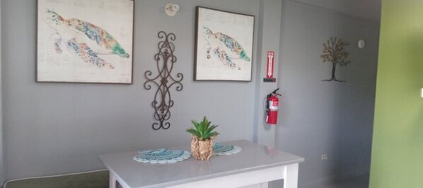 Un espace d’accueil avec un comptoir de réception blanc, deux œuvres d’art encadrées accrochées au mur, un élément décoratif en métal, une petite plante en pot, un extincteur et une décoration en forme d’arbre fixée au mur.