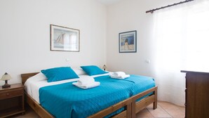 Un lit bien fait, avec une literie bleue, une tête de lit en bois et deux oreillers. On y trouve une petite table de chevet avec une lampe, et deux tableaux encadrés accrochés au mur.