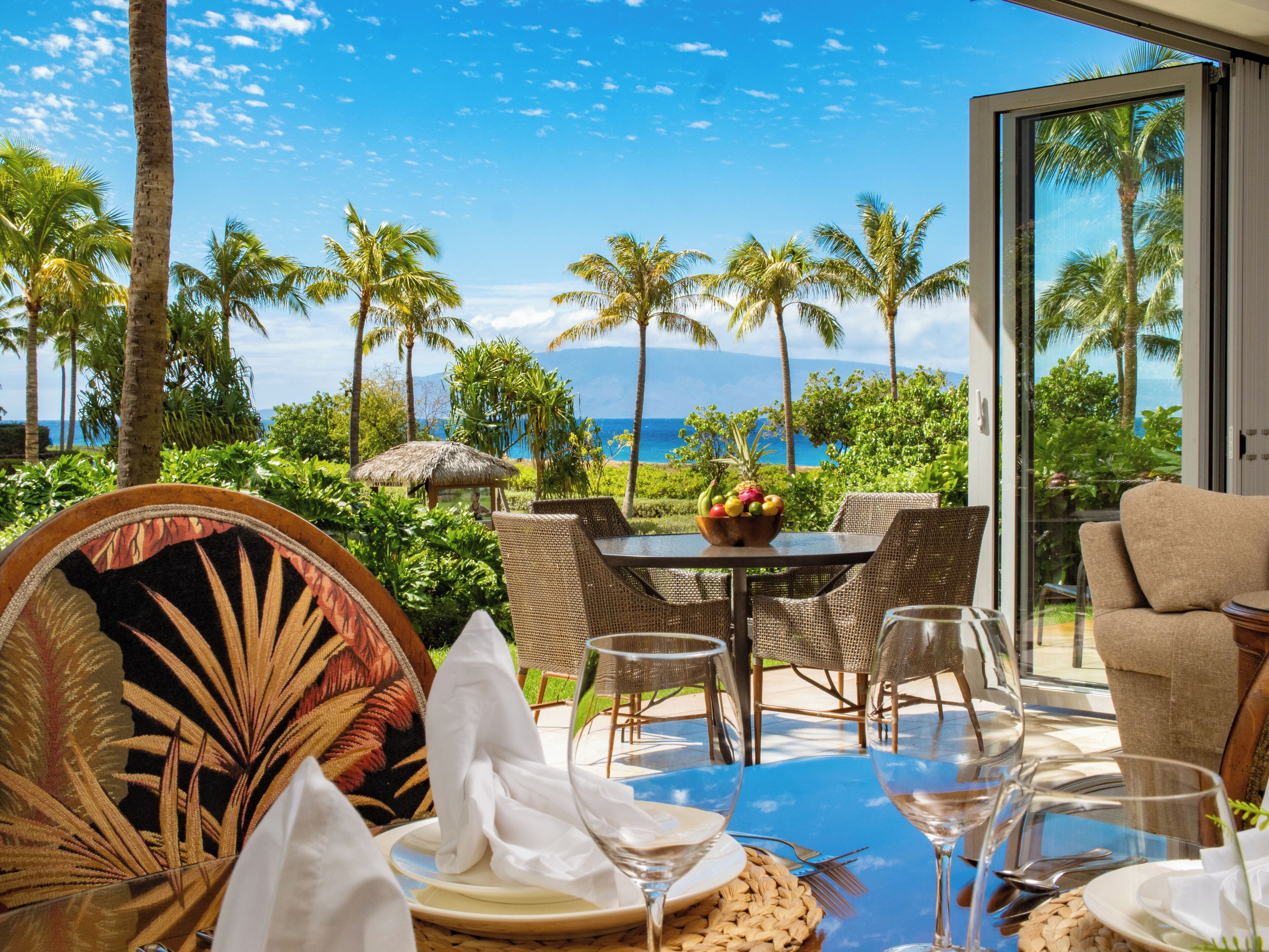 A dining area with a table set for two, overlooking a beach with palm trees and the ocean.