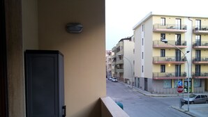 A balcony with a view of a street, a trash bin, a stop sign, and a building with balconies. - New apartment 250 m from the beach (Alghero)