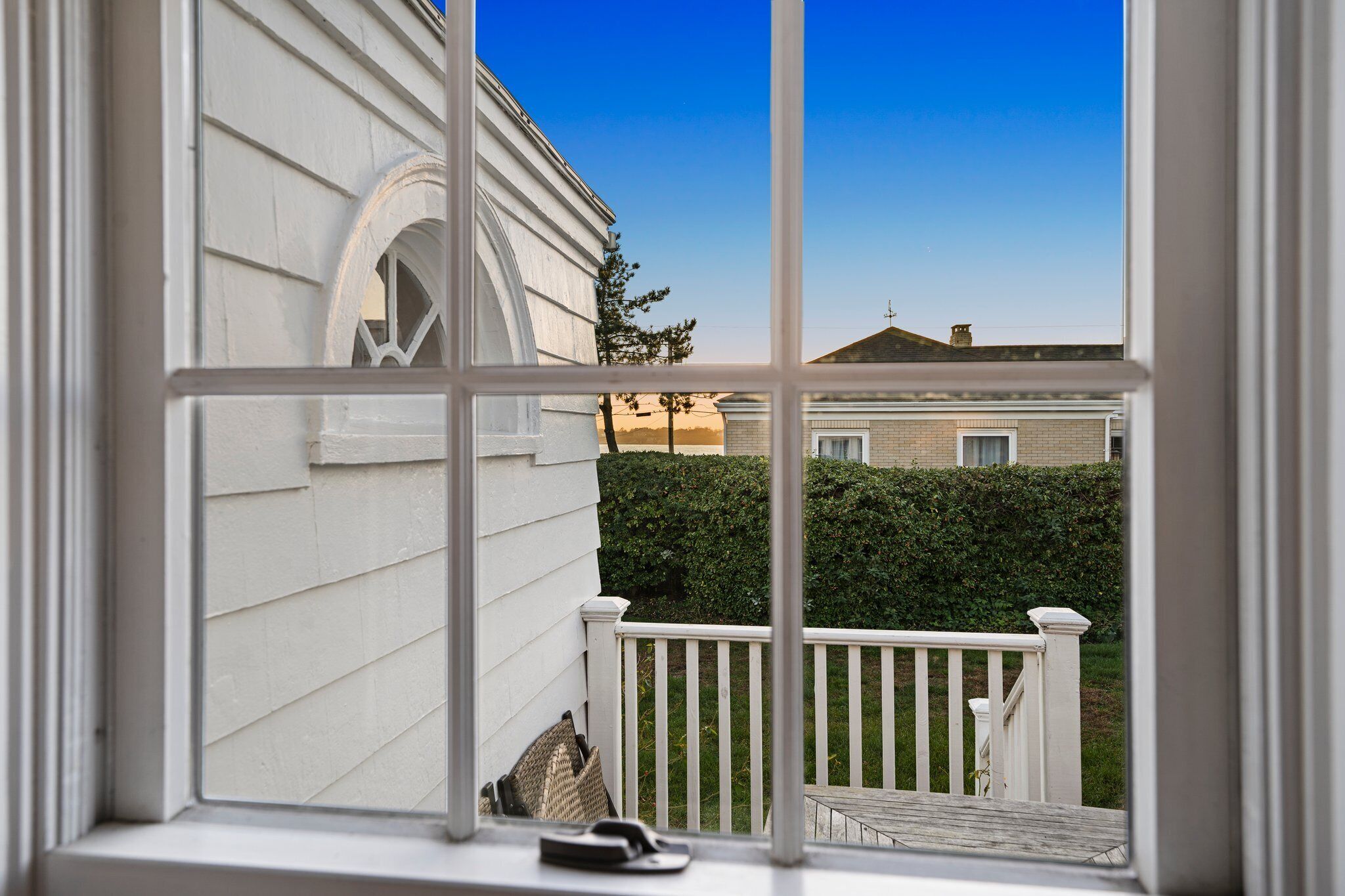 A view from a window with a white railing, a white building with a brick section, and a hedge in the background.
